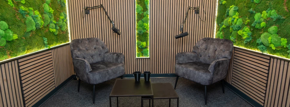 Create Lab Podcast‑Studio in Lübeck mit zwei Sesseln, Tisch und Mikrofonen vor akustisch gestalteter Wand mit Holzlamellen und Moos.