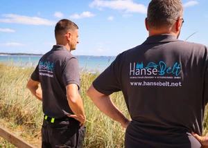 Zwei Männer in Firmen-Poloshirts der Hanse-Belt GmbH stehen in den Dünen und blicken auf das Meer an einem sonnigen Tag.
