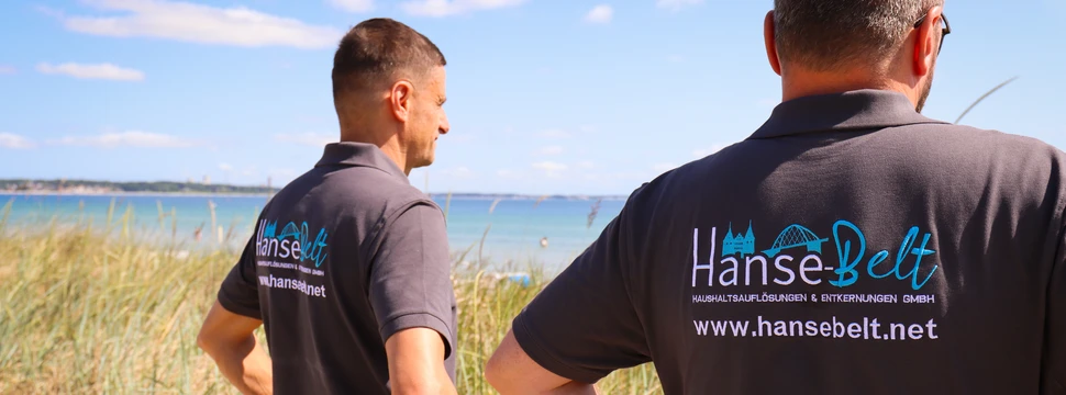 Zwei Männer in Firmen-Poloshirts der Hanse-Belt GmbH stehen in den Dünen und blicken auf das Meer an einem sonnigen Tag.
