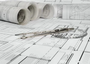 Architekturpläne und Zeichenwerkzeuge auf einem Arbeitstisch. Technische Bauzeichnungen mit Zirkel und Geodreieck, dahinter aufgerollte Architekturpläne auf weißem Papier.