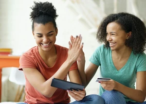 Zwei Frauen klatschen sich ab und lachen dabei. Die linke Frau hält ein Ipad in der Hand und die rechte Frau hält eine Kreditkarte