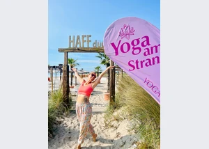 Frau steht am Strand vor den HAFFdüün bei Sonne und mit einem Schild Strandyoga YOGACOAST in Haffkrug an der Ostsee